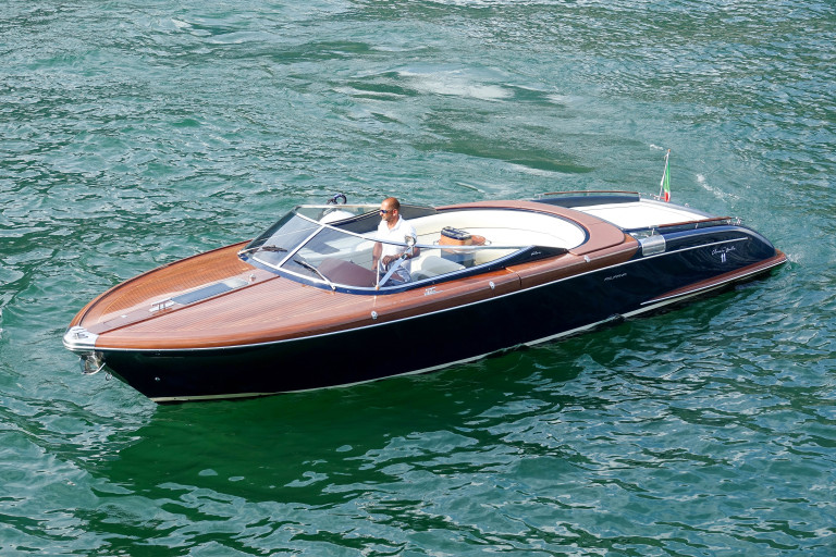 A wooden speedboat with a polished brown deck and white interior, driven by a man in a white shirt on green water.