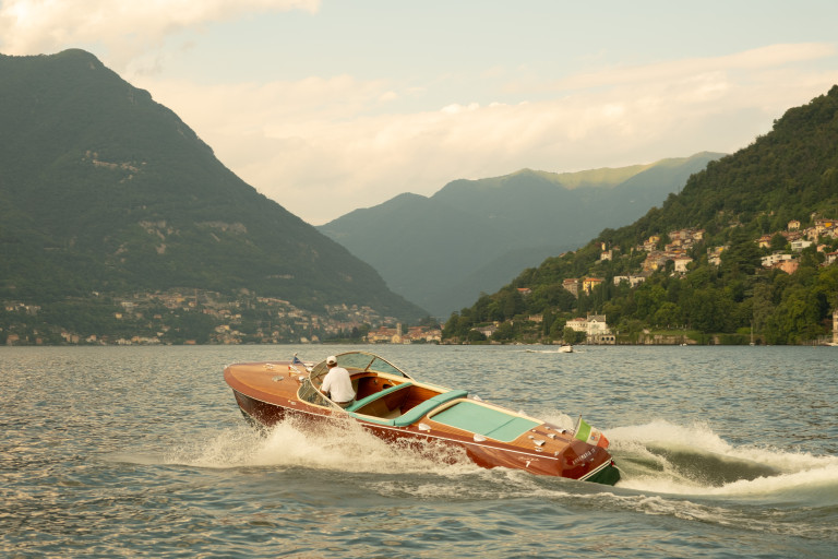 A wooden speedboat speeds across a lake with green mountains and a hillside town in the background.